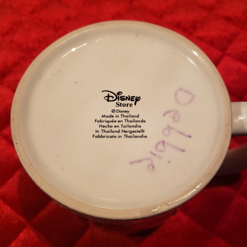 Puede incluir: Base de taza de cer&aacute;mica blanca con el logotipo de Disney Store y texto en varios idiomas. El texto incluye "Hecho en Tailandia". La palabra "Debbie" est&aacute; escrita con un rotulador morado.