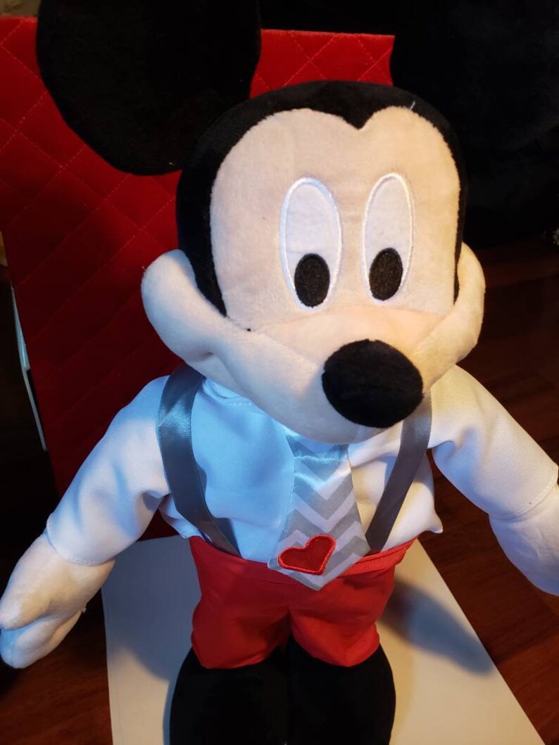 K&ouml;nnte beinhalten: Eine Mickey Mouse Pl&uuml;schpuppe mit wei&szlig;em Hemd, roter Hose und grauer Krawatte mit rotem Herz. Die Puppe hat schwarze Ohren, eine schwarze Nase und wei&szlig;e Gesichtsz&uuml;ge. Ein klassischer Disney-Charakter.
