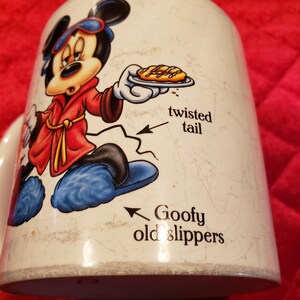 Puede incluir: Taza de cer&aacute;mica blanca con un dibujo de Mickey Mouse con una bata roja y zapatillas azules. Mickey sostiene un plato con un pastel. La taza tiene el texto "twisted tail" y "Goofy old slippers".