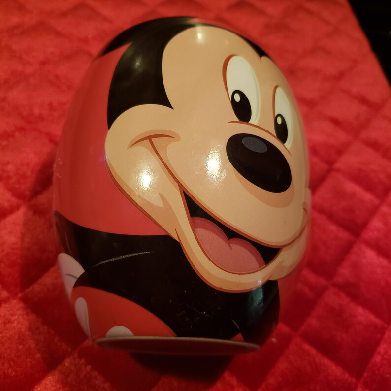 May include: A red, black, and tan Mickey Mouse-themed egg-shaped collectible. The smiling face of Mickey Mouse is prominently displayed on the front. The egg has a glossy finish and is set against a red background.