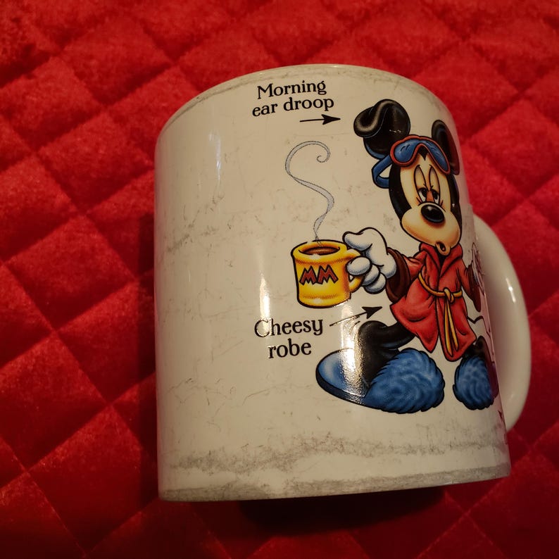 Puede incluir: Taza de cer&aacute;mica blanca con un personaje de dibujos animados de Mickey Mouse con una bata roja, sosteniendo una taza amarilla con vapor. La taza tiene el texto "Morning ear droop" y "Cheesy robe".