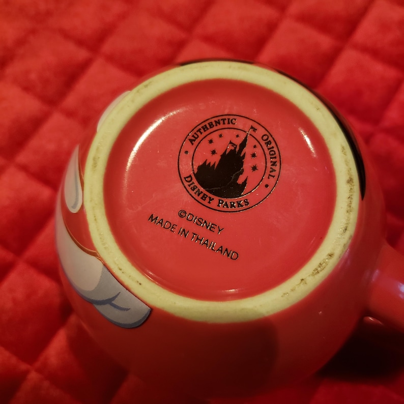 May include: Red ceramic mug with a white handle and a red interior. The bottom of the mug features a circular logo with the text "AUTHENTIC DISNEY PARKS ORIGINAL" and the text "MADE IN THAILAND".