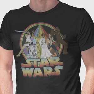 Puede incluir: Una camiseta negra de manga corta con un estampado gr&aacute;fico de personajes de Star Wars, incluyendo a Luke Skywalker, R2-D2, C-3PO, la Princesa Leia, Han Solo y Chewbacca. El dise&ntilde;o incluye un fondo circular de colores arco&iacute;ris y el logo de Star Wars.