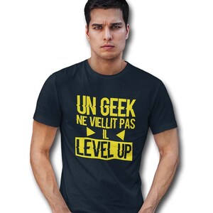 Op de afbeelding: Een donkerblauw t-shirt met gele tekst die "Un Geek Ne Vieillit Pas Il Level Up" zegt.