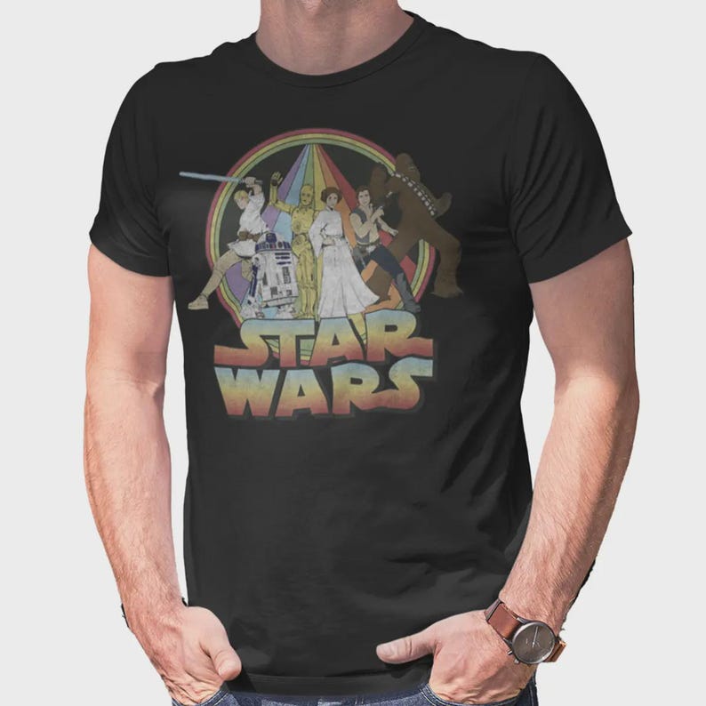 Puede incluir: Una camiseta negra de manga corta con un estampado gr&aacute;fico de personajes de Star Wars, incluyendo a Luke Skywalker, R2-D2, C-3PO, la Princesa Leia, Han Solo y Chewbacca. El dise&ntilde;o incluye un fondo circular de colores arco&iacute;ris y el logo de Star Wars.