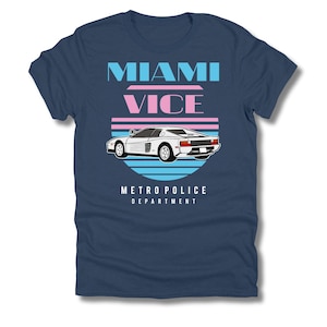 Può includere: T-shirt blu navy con grafica di un'auto sportiva bianca, la scritta "MIAMI VICE" in blu e rosa e "METRO POLICE DEPARTMENT" in bianco. Il design include un tramonto retrò e un'auto.