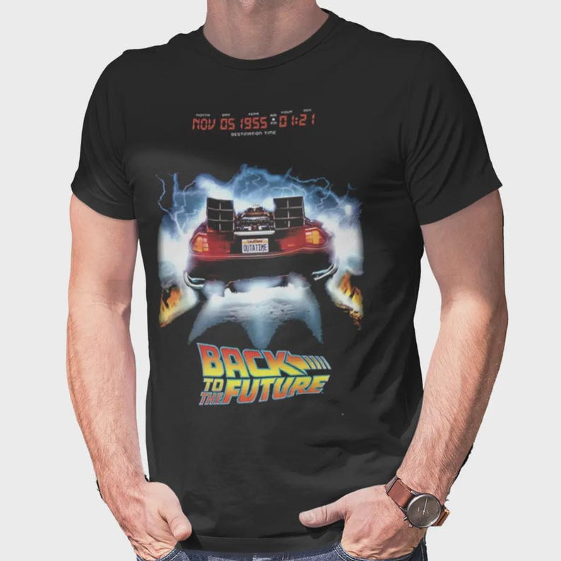 Puede incluir: Una camiseta negra de manga corta con un gr&aacute;fico de un coche deportivo rodeado de rayos azules y llamas. Sobre el coche, un texto digital rojo dice NOV 05 1955 01:21. Debajo del coche, el logotipo de Back to the Future aparece en amarillo y azul.