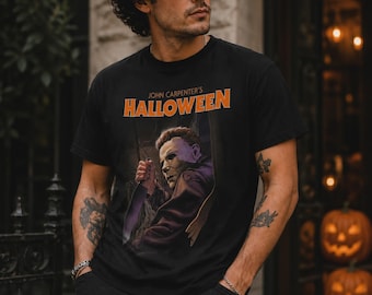 Könnte beinhalten: Ein schwarzes kurzärmeliges T-Shirt mit einem Grafikdruck der Figur Michael Myers aus dem Film John Carpenter's Halloween. Das Design enthält den orangefarbenen Schriftzug John Carpenter's Halloween über der Figur mit einem Messer.
