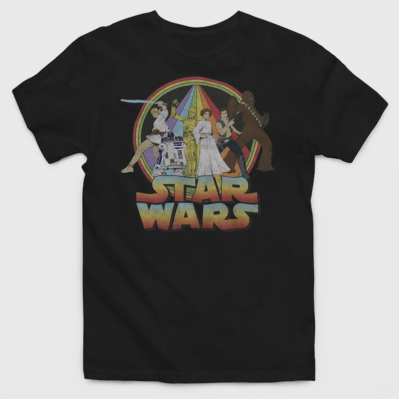 Puede incluir: Una camiseta negra de manga corta con un estampado gr&aacute;fico de personajes de Star Wars, incluyendo a Luke Skywalker, R2-D2, C-3PO, la Princesa Leia, Han Solo y Chewbacca. El dise&ntilde;o incluye un fondo circular de colores arco&iacute;ris y el texto STAR WARS.