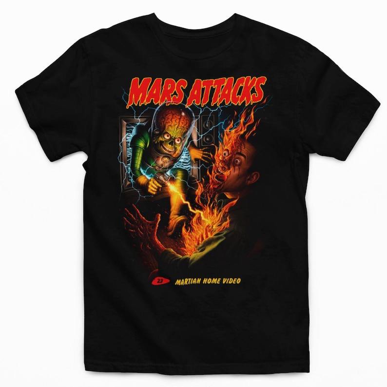 May include: A black short-sleeve t-shirt featuring a graphic print with the text MARS ATTACKS at the top and MARTIAN HOME VIDEO at the bottom. The image depicts a green alien with an exposed brain emerging from a television screen firing a ray gun.