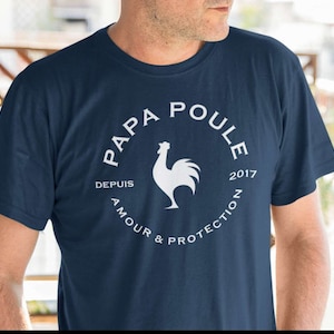 Puede incluir: Una camiseta azul marino con un gráfico blanco de un gallo y el texto "Papa Poule" en un círculo. El texto "Depuis 2017" y "Amour & Protection" también están incluidos en el gráfico.