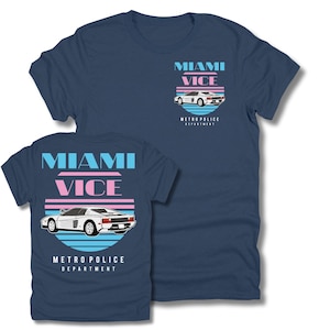Può includere: T-shirt blu navy con grafica di un'auto sportiva bianca e la scritta "Miami Vice" in blu e rosa. La maglietta include anche la scritta "Metro Police Department". Il disegno è ripetuto sul retro della maglietta.