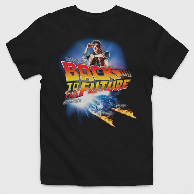 May include: A black short-sleeved t-shirt featuring a graphic print of the Back to the Future movie logo, a character wearing goggles and a vest, and a car leaving trails of fire. The design includes the text Steven Spielberg and Robert Zemeckis.