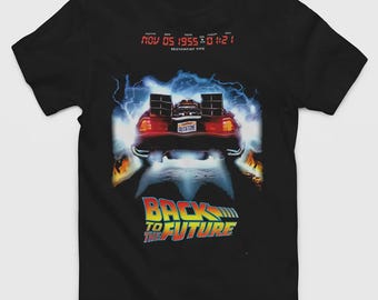 Könnte beinhalten: Ein schwarzes T-Shirt mit einer Grafik eines Sportwagens mit einem Nummernschild, auf dem OUTATIME steht, umgeben von blauen Blitzen und Flammen. Über dem Auto steht in roter Digitalanzeige NOV 05 1955 01:21. Darunter steht BACK TO THE FUTURE.