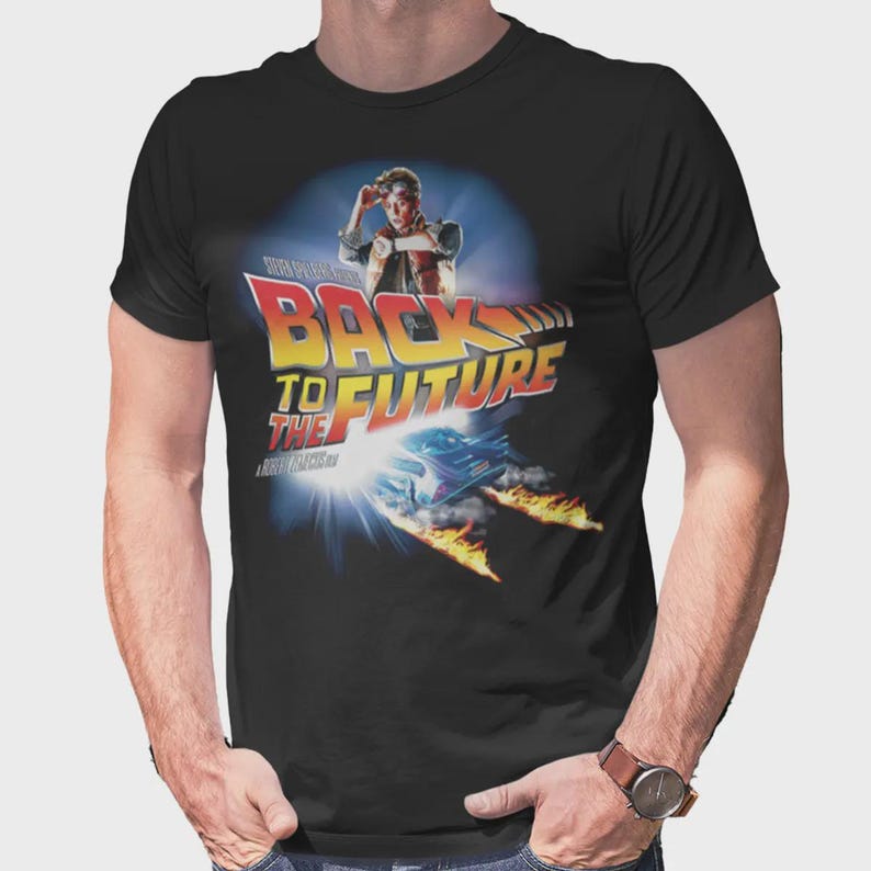 May include: A black short-sleeve t-shirt featuring a graphic print of the Back to the Future movie logo, a character looking at a watch, and a car leaving fire trails. The design includes the text Back to the Future in red and yellow.