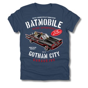 Puede incluir: Camiseta azul marino con un gráfico del Batmóvil, un coche negro con detalles rojos, y el texto "Batmobile" y "Gotham City Garage Inc." La camiseta también incluye el texto "To the Batcave!!".