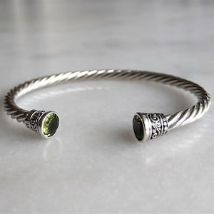 Könnte beinhalten: Ein silbernes, gedrehtes Armreif-Armband mit zwei grünen Edelstein-Akzenten.