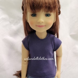 May include: A doll wearing a purple short-sleeved shirt and blue jeans. The doll has long brown hair with a braid on each side of her head. The doll has blue eyes and a light complexion. The doll is standing on a white surface. The text "urbandollclothes.com" is visible on the shirt.