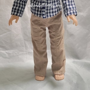 May include: A doll wearing a blue and white checkered shirt and khaki pants. The doll has blonde hair and blue eyes. The shirt has a button-up front and long sleeves. The pants have a straight leg and are made of a soft, comfortable fabric. The doll is standing on a white background.  "urbandollclothes.com" is printed on the shirt.