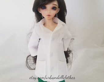 May include: A white lab coat with a pocket and buttons, worn by a doll with long brown hair and green pants. The doll is wearing a white lab coat with a pocket and buttons. The doll has long brown hair and is wearing green pants. The doll is standing on a white background. etsy.com/urbandollclothes