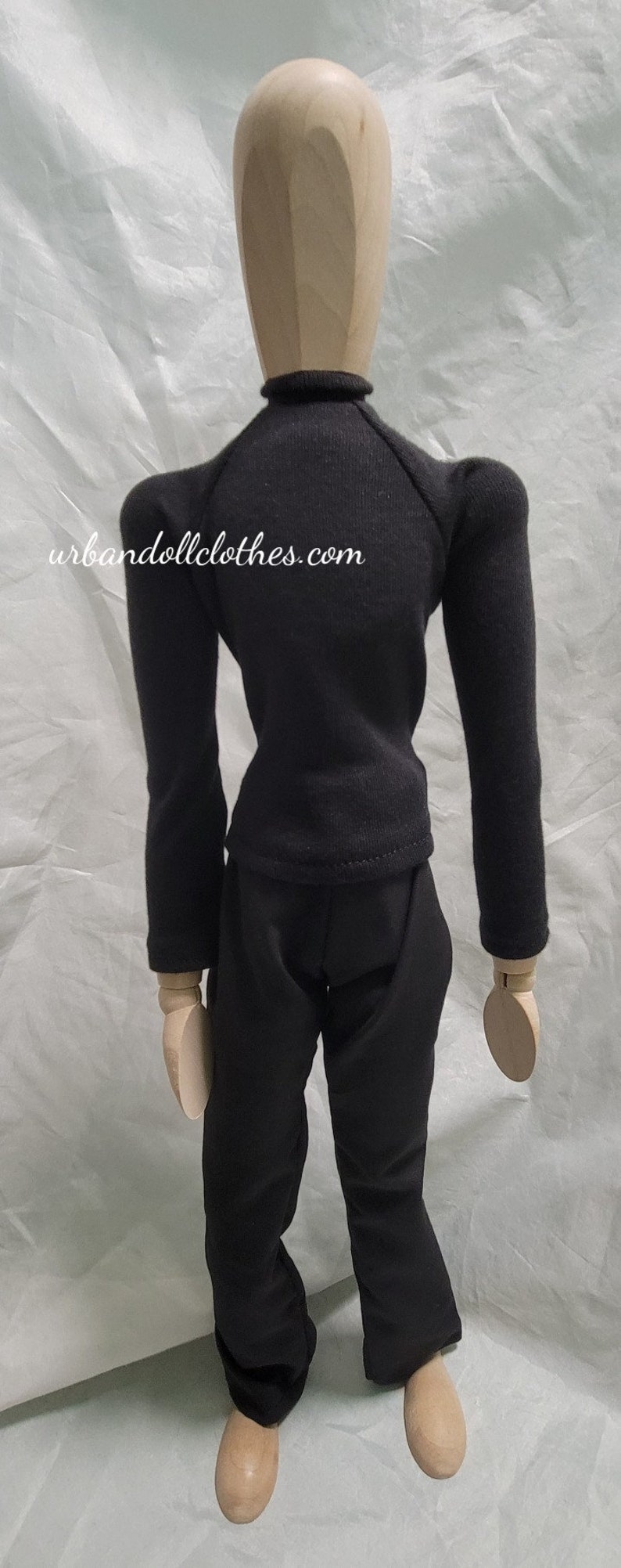 Op de afbeelding: Een zwarte poppenoutfit bestaande uit een top met lange mouwen en een broek. De top heeft een col. De website urbandollclothes.com staat op de achterkant van de top. De outfit wordt getoond op een houten mannequin.
