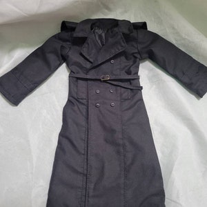 Op de afbeelding: Een zwarte trenchcoat met een dubbele rij knopen, een riem met een goudkleurige gesp en een reverskraag. De jas heeft lange mouwen en is gemaakt van een gladde stof. De jas is ontworpen voor een pop.