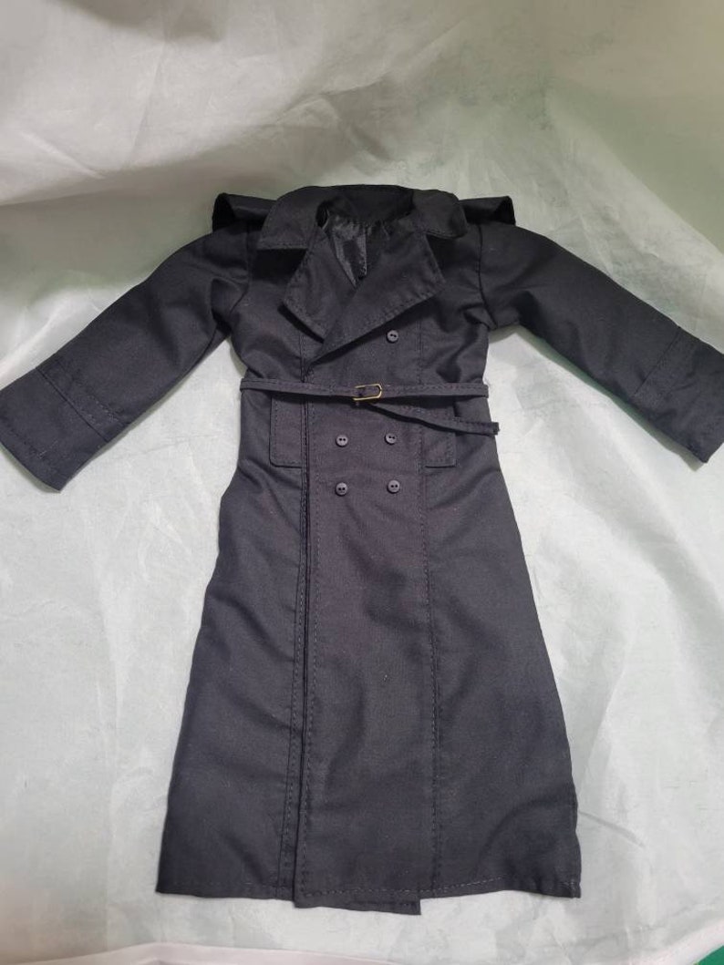 Op de afbeelding: Een zwarte trenchcoat met een dubbele rij knopen, een riem met een goudkleurige gesp en een reverskraag. De jas heeft lange mouwen en is gemaakt van een gladde stof. De jas is ontworpen voor een pop.