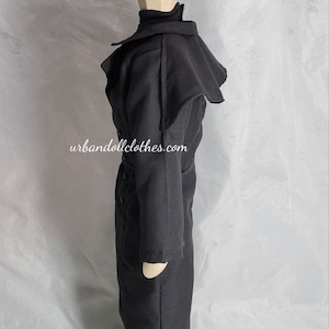 Op de afbeelding: Een zwarte jas voor poppen met een hoge kraag en een cape-achtig detail op de schouders. De jas heeft een knoopsluiting aan de voorkant en een riem in de taille. De laarzen hebben zilverkleurige knopen. De website urbandollclothes.com staat op de jas.