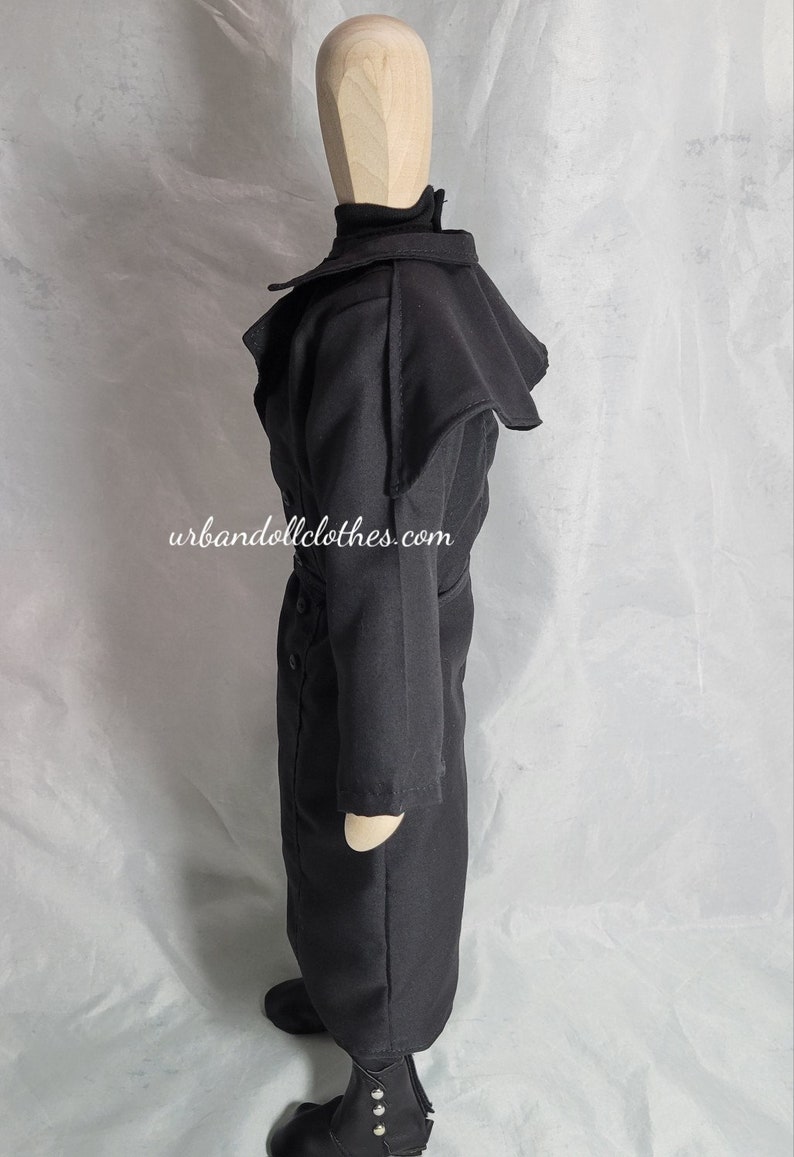 Op de afbeelding: Een zwarte jas voor poppen met een hoge kraag en een cape-achtig detail op de schouders. De jas heeft een knoopsluiting aan de voorkant en een riem in de taille. De laarzen hebben zilverkleurige knopen. De website urbandollclothes.com staat op de jas.
