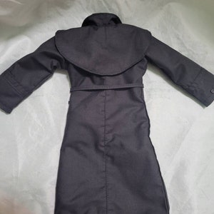 Op de afbeelding: Een zwarte trenchcoat met een brede kraag en een riem in de taille. De jas heeft lange mouwen en een knoopdetail op de manchetten. De jas is gemaakt van een gladde, donkere stof.
