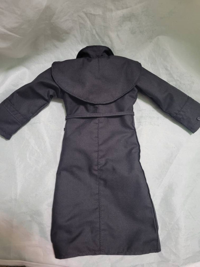 Op de afbeelding: Een zwarte trenchcoat met een brede kraag en een riem in de taille. De jas heeft lange mouwen en een knoopdetail op de manchetten. De jas is gemaakt van een gladde, donkere stof.