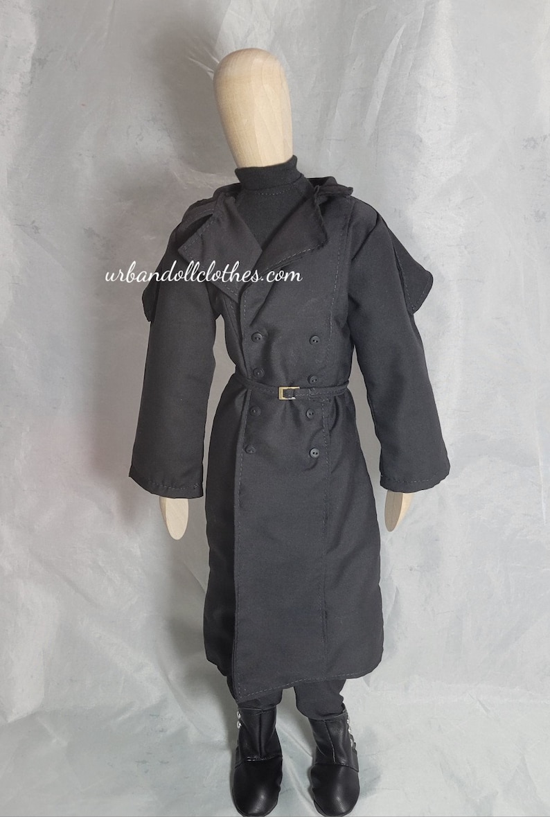 Op de afbeelding: Een zwarte trenchcoat met een dubbele rij knopen, een riem en een hoge kraag. De jas wordt gedragen over een zwarte coltrui en broek, gecombineerd met zwarte laarzen. De website urbandollclothes.com is zichtbaar.