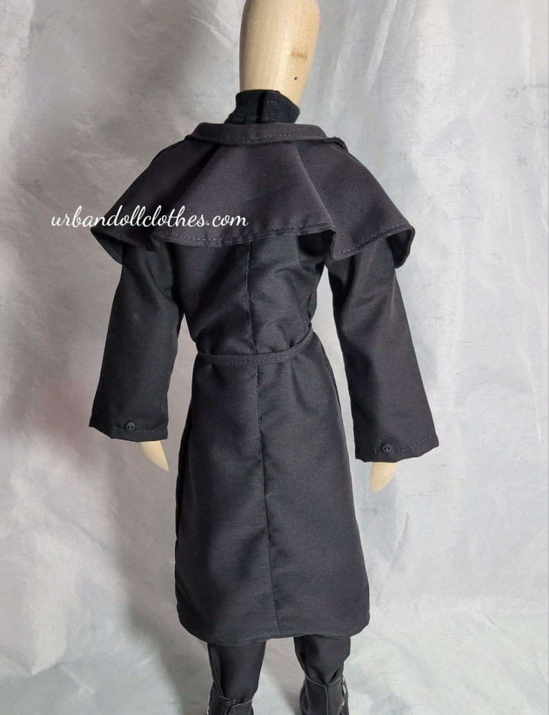 Op de afbeelding: Een zwarte poppenjas met een cape-achtige kraag, lange mouwen en een riem. De jas is dichtgeknoopt en wordt over een zwarte coltrui gedragen. De website urbandollclothes.com staat op de achterkant van de jas.