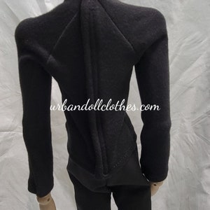 Op de afbeelding: Een zwart poppenoutfit, bestaande uit een top met lange mouwen en een broek, gepresenteerd op een houten mannequin. De top heeft een hoge kraag en een ritssluiting aan de achterkant. De broek is uitlopend. De website urbandollclothes.com is zichtbaar.