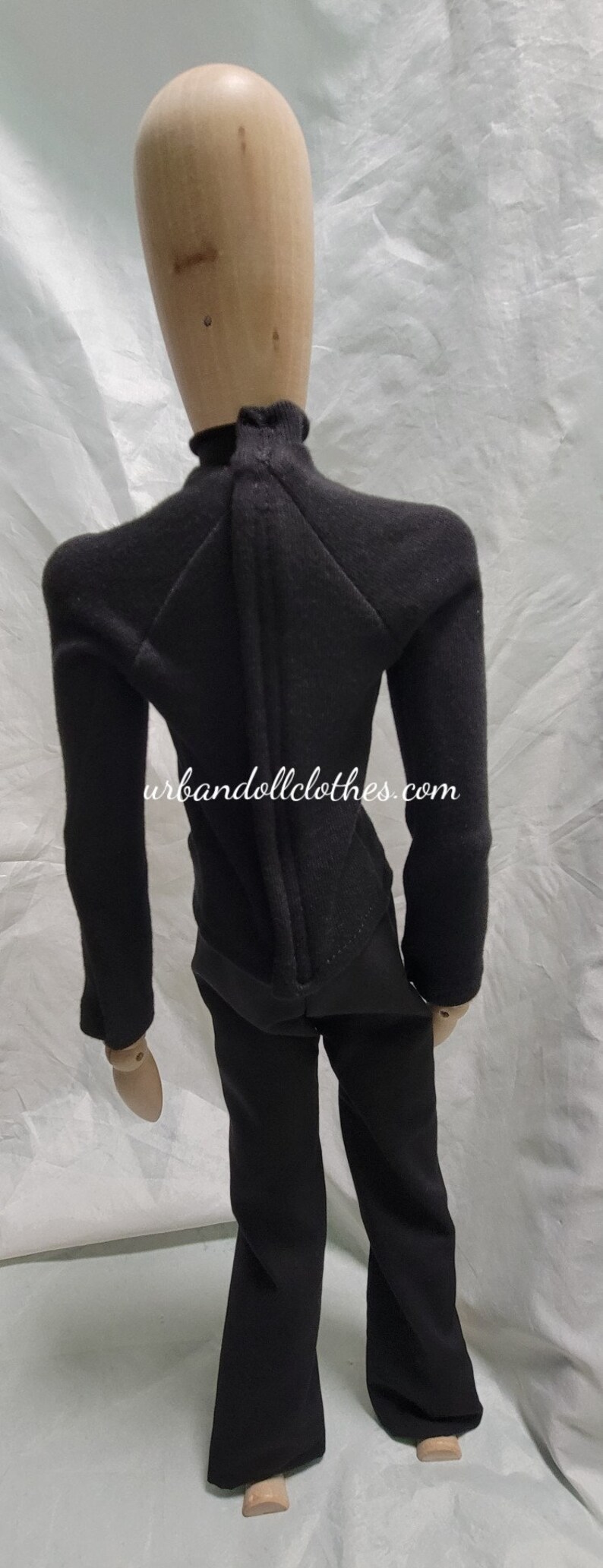 Op de afbeelding: Een zwart poppenoutfit, bestaande uit een top met lange mouwen en een broek, gepresenteerd op een houten mannequin. De top heeft een hoge kraag en een ritssluiting aan de achterkant. De broek is uitlopend. De website urbandollclothes.com is zichtbaar.