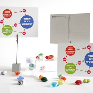Puede incluir: Dos tarjetas blancas con un diagrama de flujo rojo, verde y azul en cada una. El diagrama de flujo pregunta: "¿Tienes un problema en la vida?" y "¿Puedes hacer algo al respecto?" Si la respuesta a ambas preguntas es sí, entonces el diagrama de flujo dice: "Entonces, ¿por qué preocuparse?" Las tarjetas están sobre una superficie blanca con una caja de acrílico transparente y canicas de vidrio de colores.