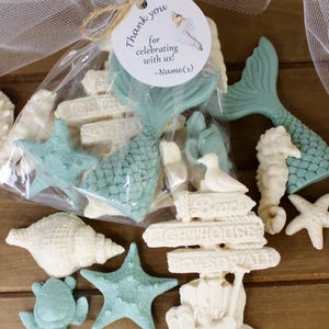 May include: A collection of ocean-themed party favors, including starfish, seashells, mermaid tails, and a seahorse. Some items are light blue and white. A clear bag contains favors with a tag that reads "Thank you for celebrating with us!"