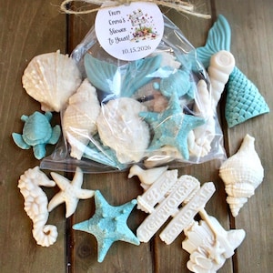 May include: A clear plastic bag filled with assorted white and light blue ocean-themed soaps shaped like seashells, starfish, sea turtles, and mermaid tails. A round tag attached with twine reads From Emma's Shower to Yours! 10.15.2026.