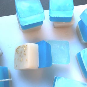 May include: A collection of square-shaped soap bars featuring a layered design. Each bar consists of a translucent bright blue top section and an opaque cream-colored bottom section. One bar displays small, light brown flecks embedded in the cream base.