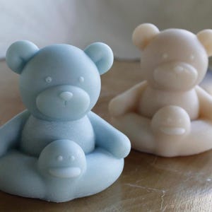 May include: Two bear-shaped figures, one light blue and one cream-colored, each featuring a smaller circular element at the base. The figures have rounded ears, defined snouts, and simple facial features, presented as solid, smooth-textured objects.