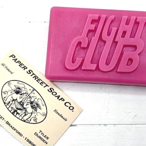 May include: A pink bar of soap with the words "FIGHT CLUB" embossed on the top. A business card from Paper Street Soap Co. is in the foreground. The card has an illustration of cherubs and the text "All Natural" and "Handmade".