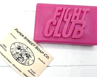 Könnte beinhalten: Ein rosa Seifenstück mit den Worten "FIGHT CLUB" auf der Oberseite. Eine Visitenkarte von Paper Street Soap Co. befindet sich im Vordergrund. Die Karte hat eine Illustration von Engeln und den Text "All Natural" und "Handmade".
