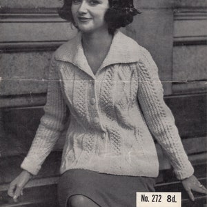 May include: A white knitted cardigan with a button fastening and a collar. The cardigan is being modelled by a woman with dark hair and a black hat. The text "“ARAN” DOUBLE KNITTING" and "No. 272 8d." is visible in the image.