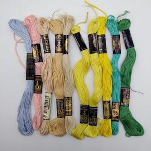 Könnte beinhalten: Sortiment von zehn Knäuel Anchor-Stickgarn in verschiedenen Farben, darunter hellblau, rosa, beige, gelb und grün. Jedes Knäuel hat ein Etikett mit dem Markennamen, der Farbnummer und dem Aufdruck "Fast Colour".