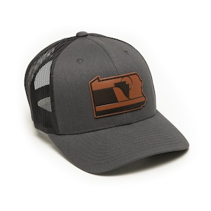 May include: A gray and black trucker hat with a brown leather patch featuring the outline of Pennsylvania. The patch has a stylized design of a sunrise over a mountain range.