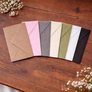 May include: A collection of colorful envelopes arranged on a wooden surface. The envelopes are in various colors, including brown, pink, gray, cream, olive green, white, and black. Delicate white flowers are scattered around the envelopes.