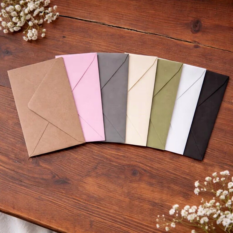 May include: A collection of colorful envelopes arranged on a wooden surface. The envelopes are in various colors, including brown, pink, gray, cream, olive green, white, and black. Delicate white flowers are scattered around the envelopes.