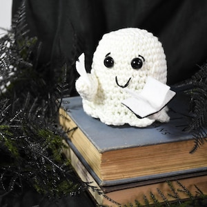 May include: A crocheted white ghost doll with black eyes and a smiling mouth, holding a small book and a piece of paper. The ghost is sitting on top of a stack of old books with yellowed pages, surrounded by black decorative foliage.