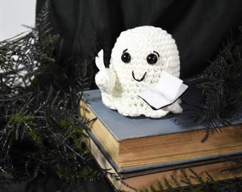 Puede incluir: Una muñeca fantasma blanca de ganchillo con ojos negros y una boca sonriente, sosteniendo un pequeño libro y un trozo de papel. El fantasma está sentado encima de una pila de libros viejos con páginas amarillentas, rodeado de follaje decorativo negro.