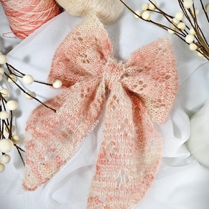 May include: A delicate, hand-knitted bow made with a soft, pink and white yarn. The bow is tied in a loose knot and has a lacy pattern. The bow is displayed on a white background.